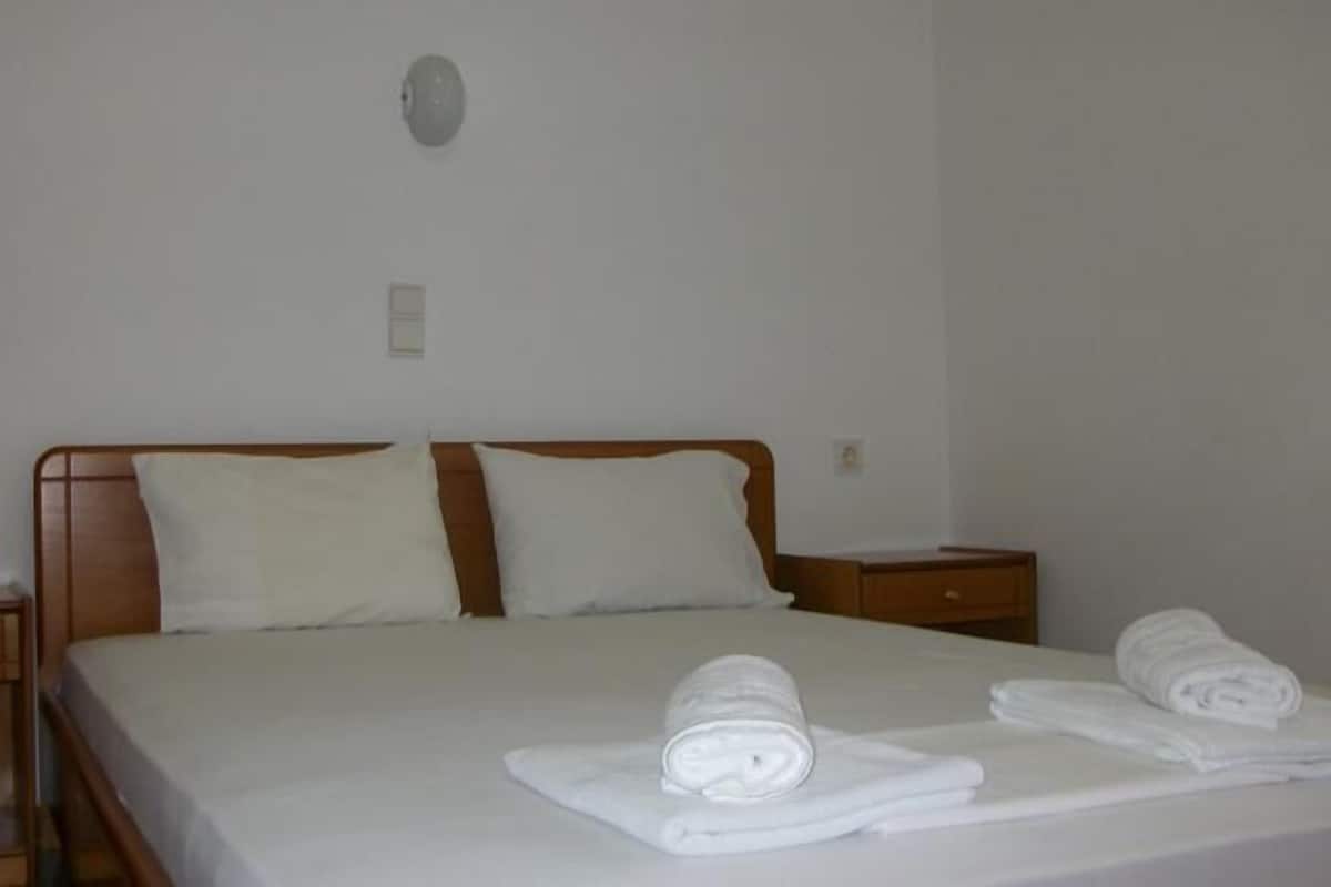 A hotel room with a bed, two pillows, a nightstand, and a rolled towel on the bed.