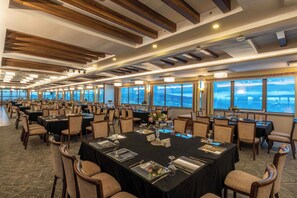 Uma ampla sala de banquetes com várias mesas postas para refeições, grandes janelas com vista e um design moderno no teto.
