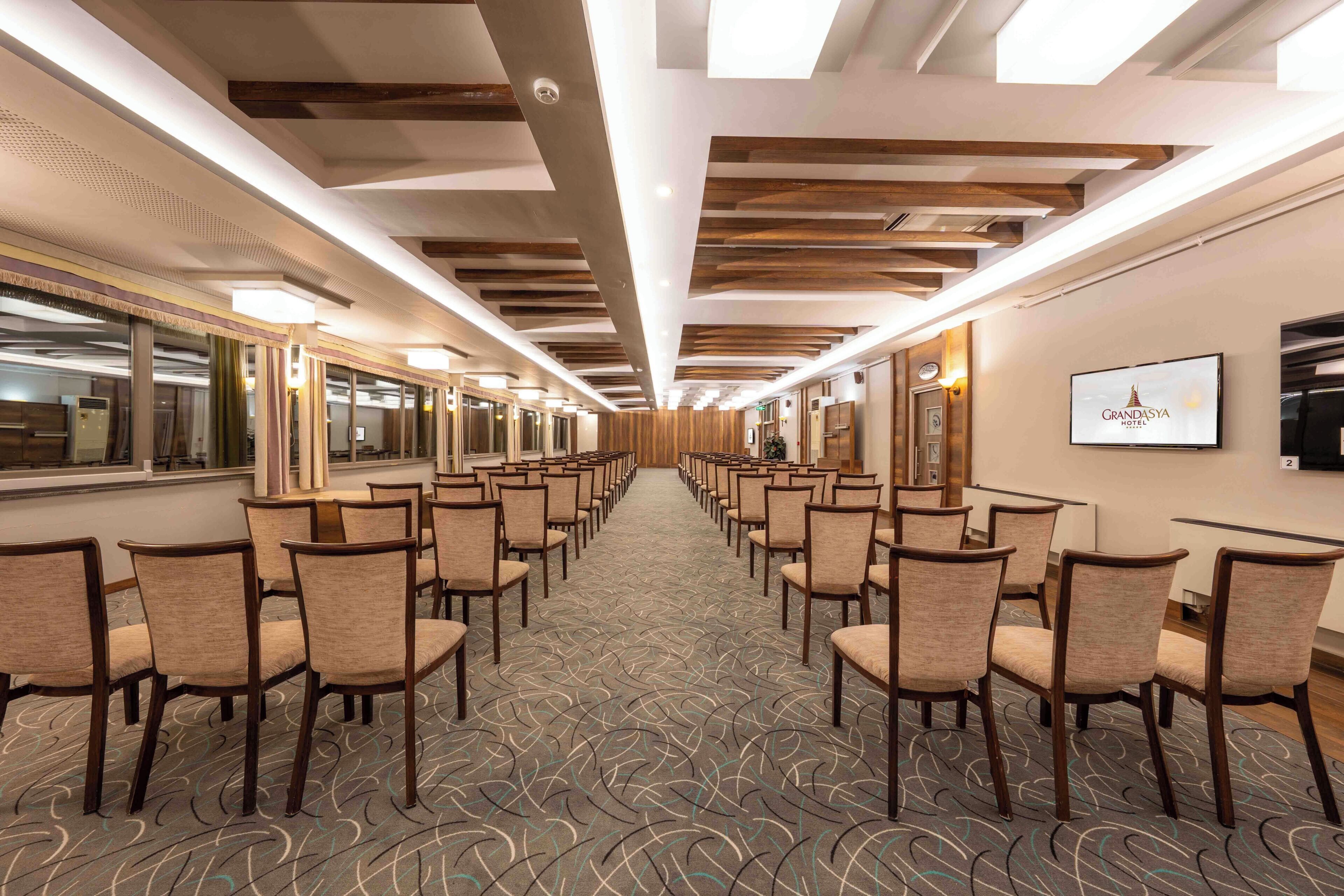 A conference room with rows of chairs, a patterned carpet, wooden ceiling beams, and a wall-mounted television.