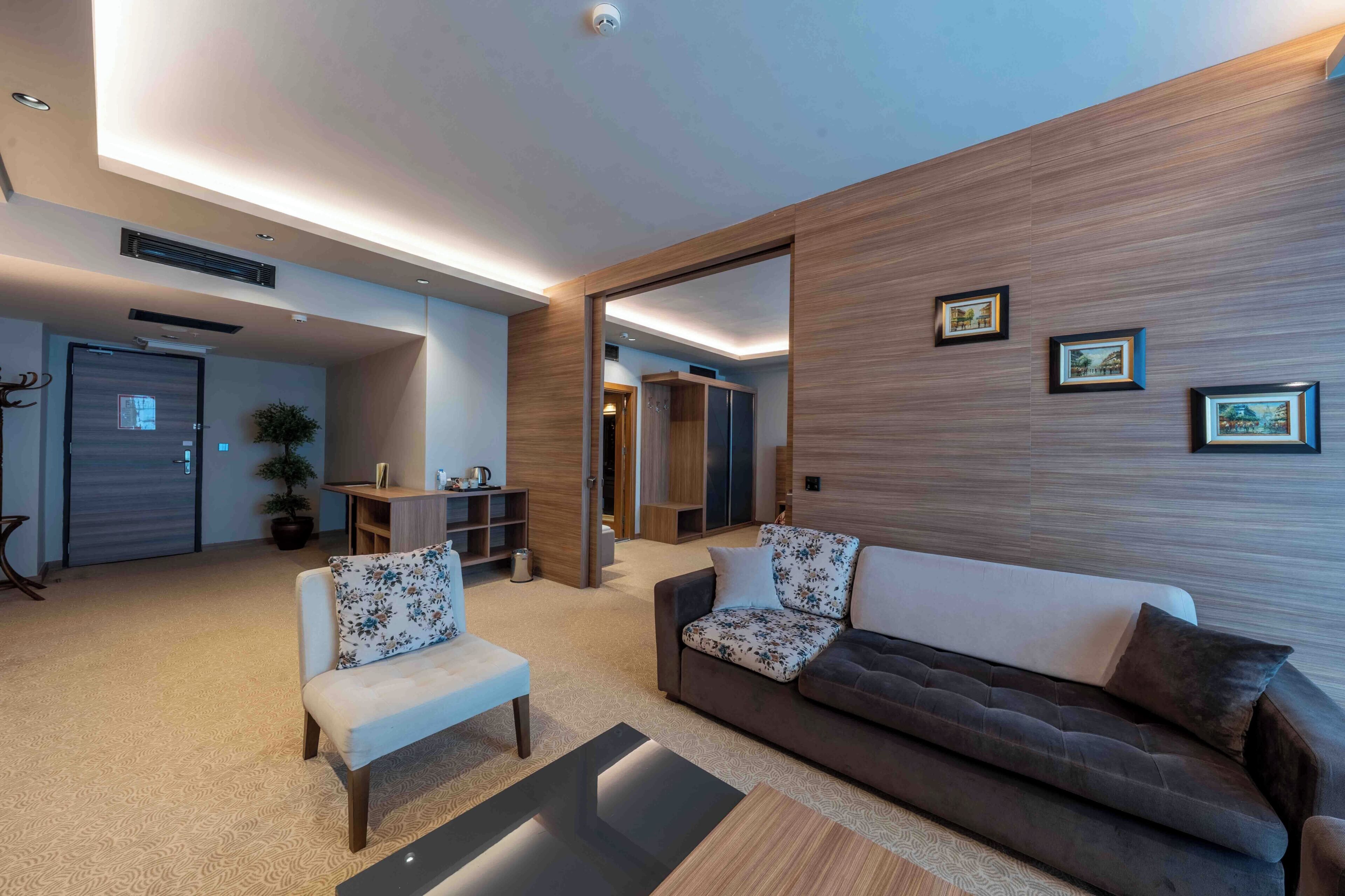 A modern living room with a sofa, armchair, and a coffee table. There is a door leading to another room, a potted plant, and a small table with a lamp.