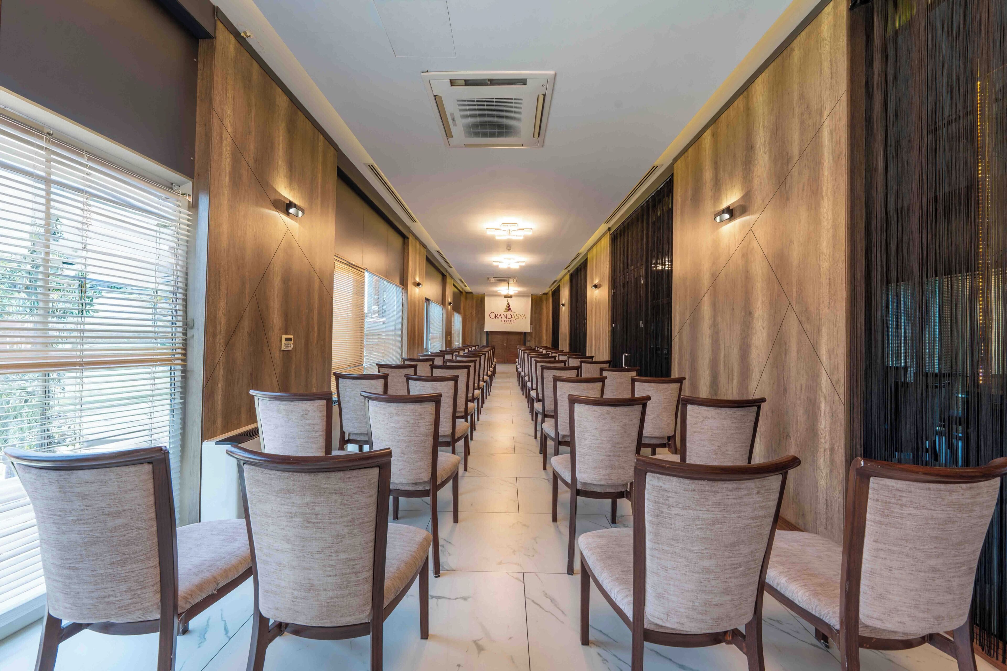 Un couloir d’hôtel bordé de rangées de chaises, avec des lambris en bois et une enseigne indiquant « GRANDOSHA ».