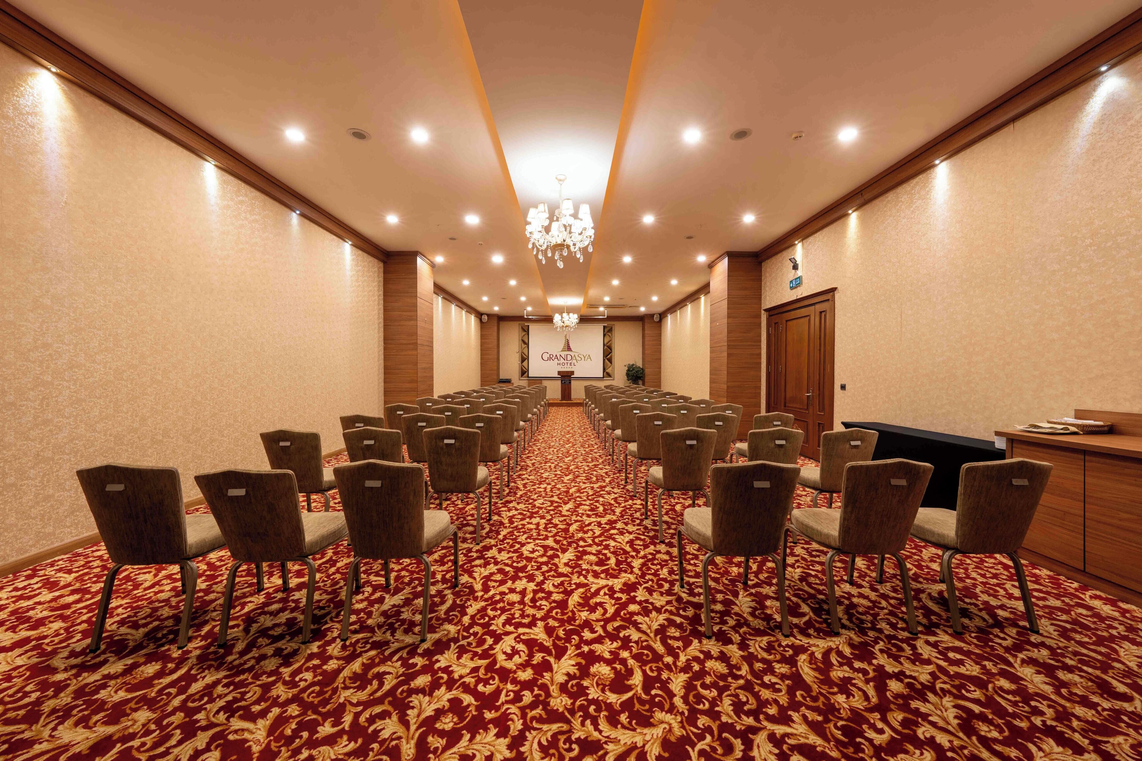 A conference room with rows of chairs, a red carpet, a chandelier, and a reception desk.