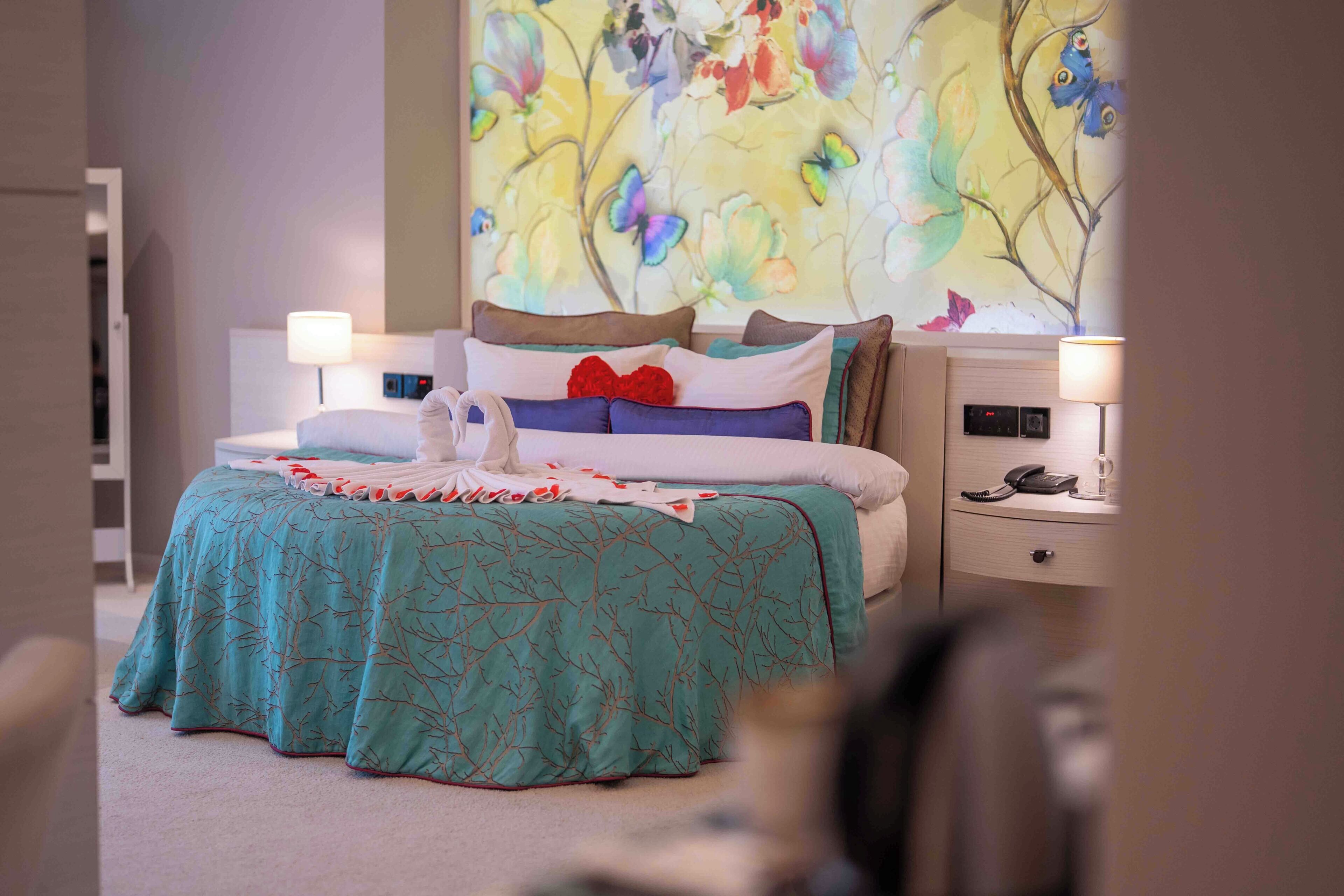 A hotel room with a bed, two bedside tables, a nightstand, a wall with a floral mural, and a door.