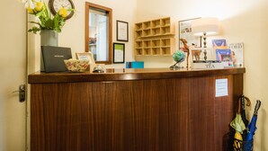A wooden reception desk with a laptop, framed certificates, a vase with flowers, a tea station, and a wall-mounted shelf with books and a globe. - George Oxford Hotel (Oxford)