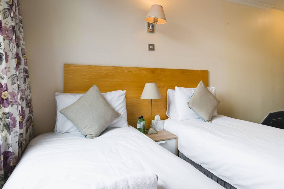 A hotel room with two beds, each with white bedding and pillows, a wooden headboard, a bedside table with a lamp, and a floral curtain.