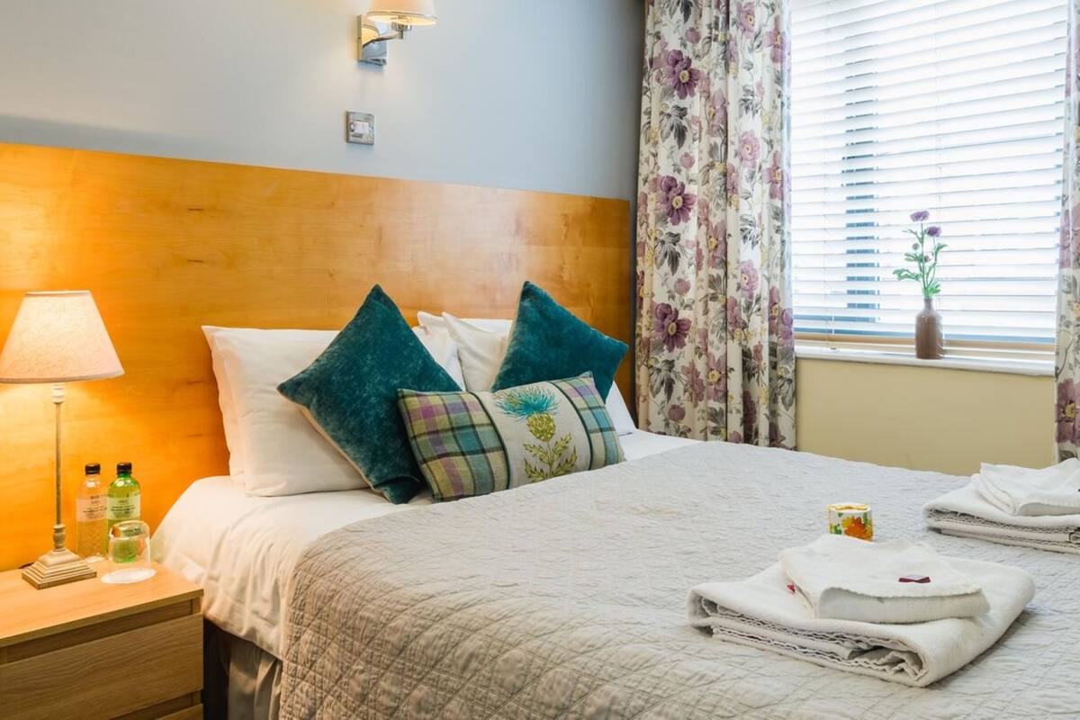 A hotel room with a wooden headboard, a bedside lamp, a nightstand with toiletries, a bed with a quilted bedspread and decorative pillows, a window with floral curtains, and a vase with a plant.
