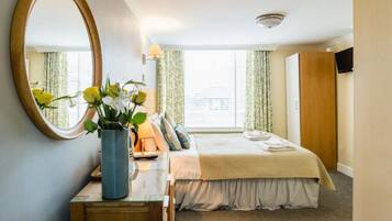 Una habitación de hotel con una cama, una mesita de noche con un florero y un espejo, y un televisor montado en la pared.