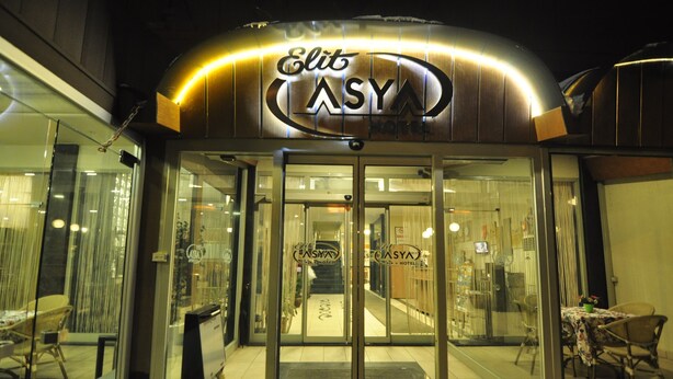 El exterior del Hotel Elite ASYA, con una entrada de vidrio, una escalera interior visible y un área de comedor con mesas y sillas afuera.