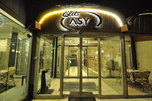 El exterior del Hotel Elite ASYA, con una entrada de vidrio, una escalera interior visible y un área de comedor con mesas y sillas afuera.