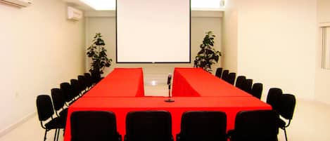 A conference room with a long table, black chairs, a red tablecloth, a projector screen, and two potted plants.