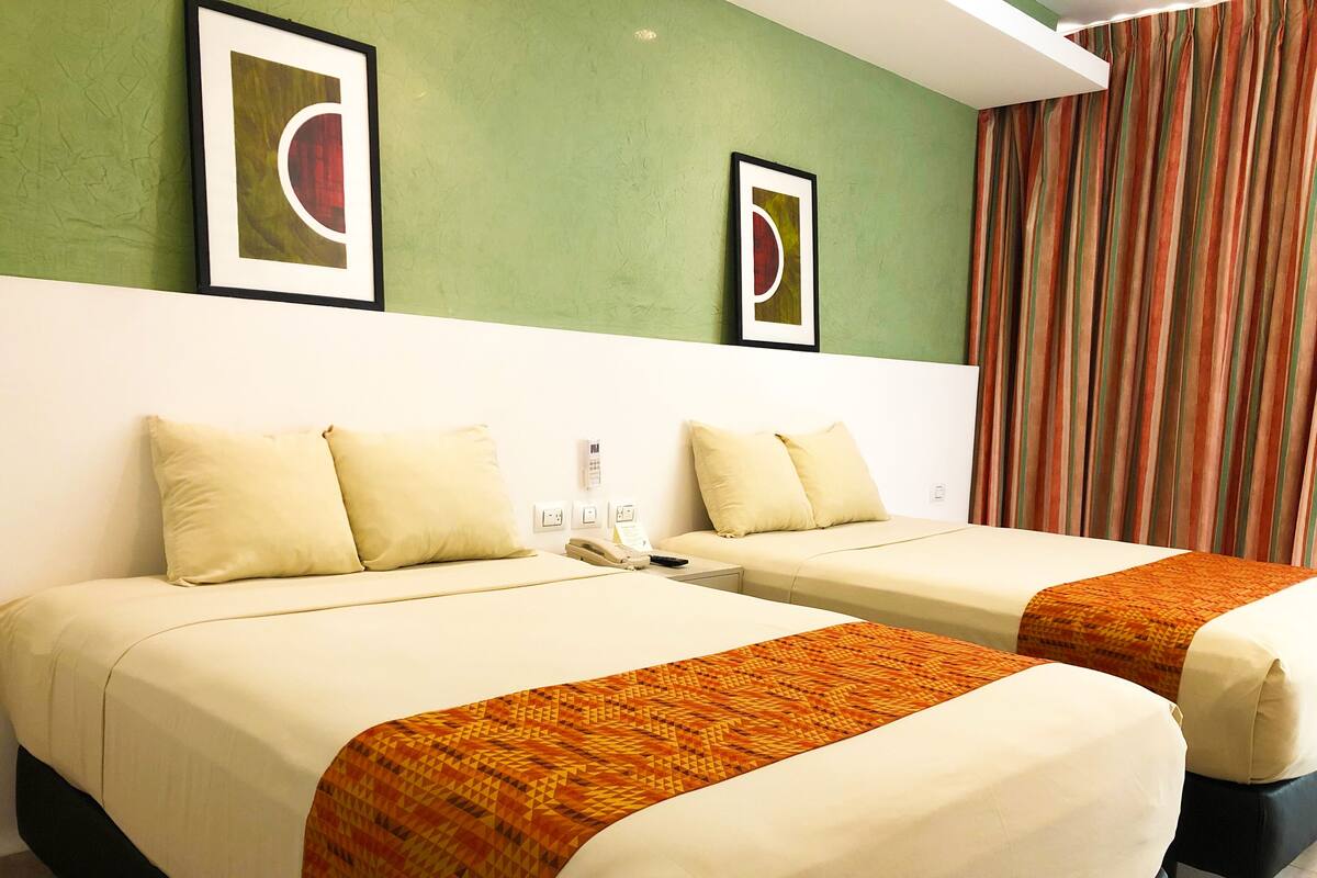 Habitación de hotel con dos camas, cada una con una colcha blanca y almohadas a juego. Se observan cuadros enmarcados en la pared, un teléfono en la mesita de noche y una cortina a rayas.