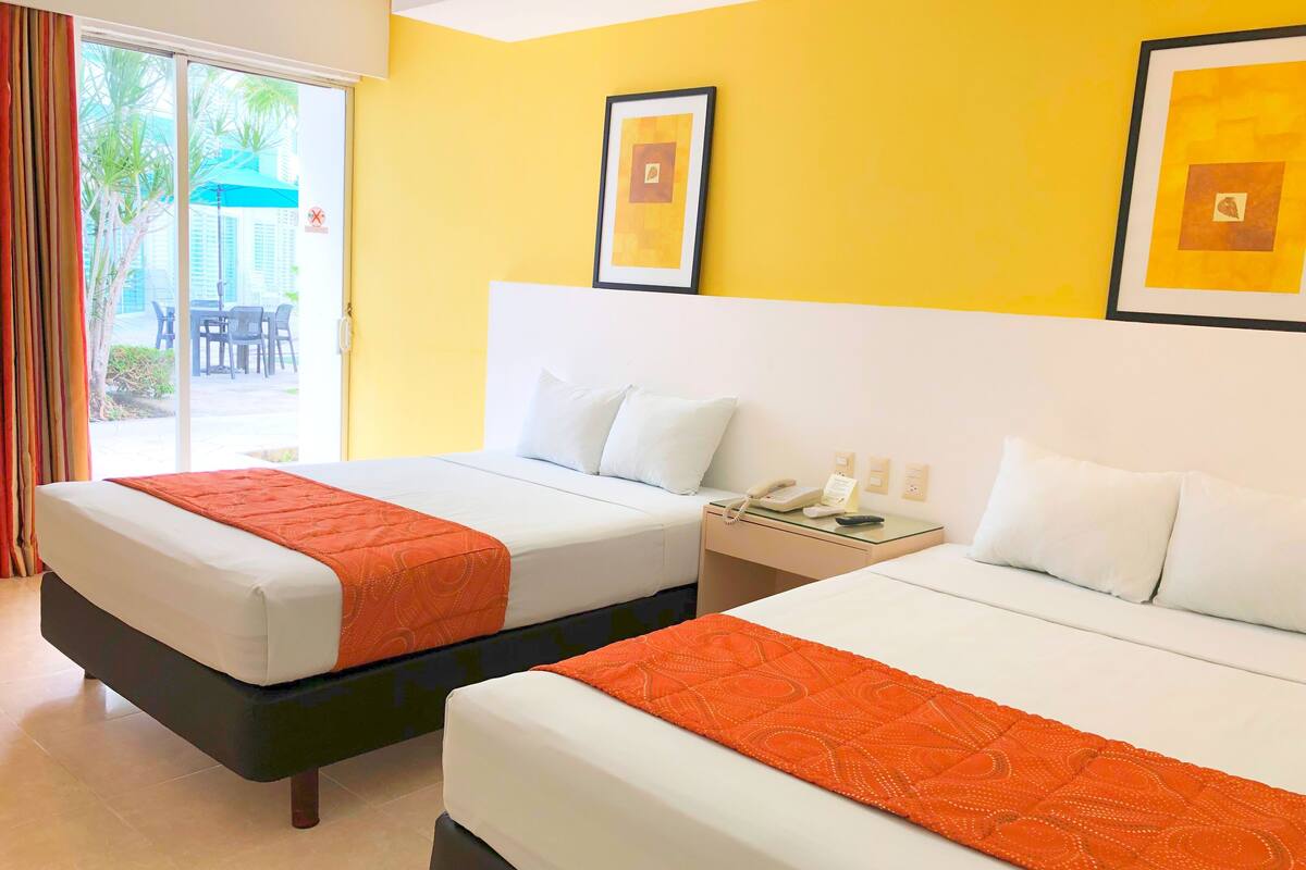 Habitación de hotel con dos camas, un ventanal de vidrio que da a un espacio exterior y una mesita de noche pequeña con un teléfono y un control remoto.
