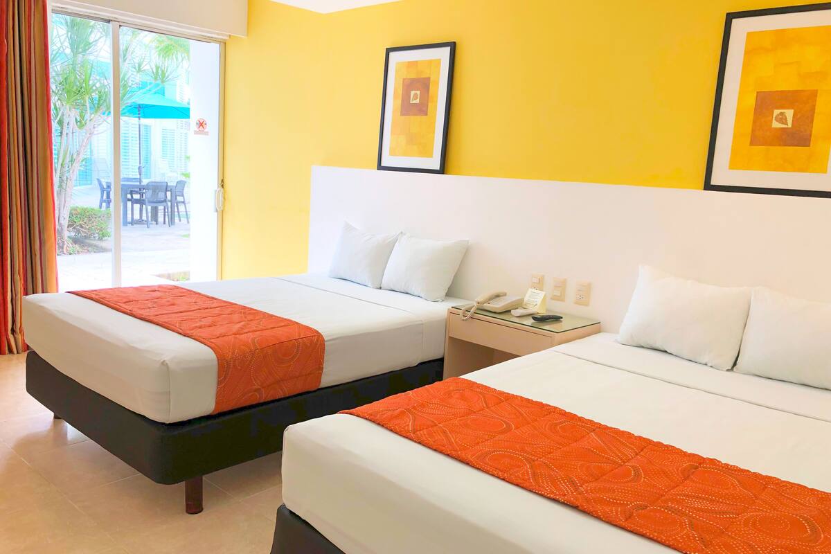 Habitación de hotel con dos camas, un vidrio que da a un área exterior y una pared amarilla con obras de arte enmarcadas.