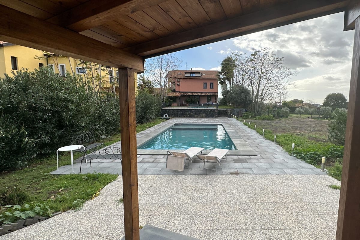 A swimming pool with a pergola overhead, surrounded by a garden and a view of a house in the background.