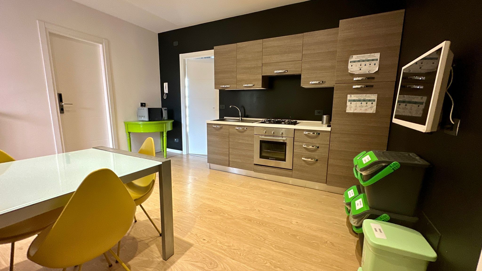 A compact kitchen with a built-in oven, a sink, and a stove. There is a dining table with chairs and a small green table with a microwave.