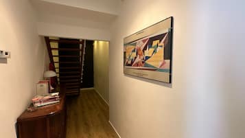 Un pasillo con una escalera de madera, un cuadro abstracto enmarcado y una pequeña mesa con libros y una lámpara.