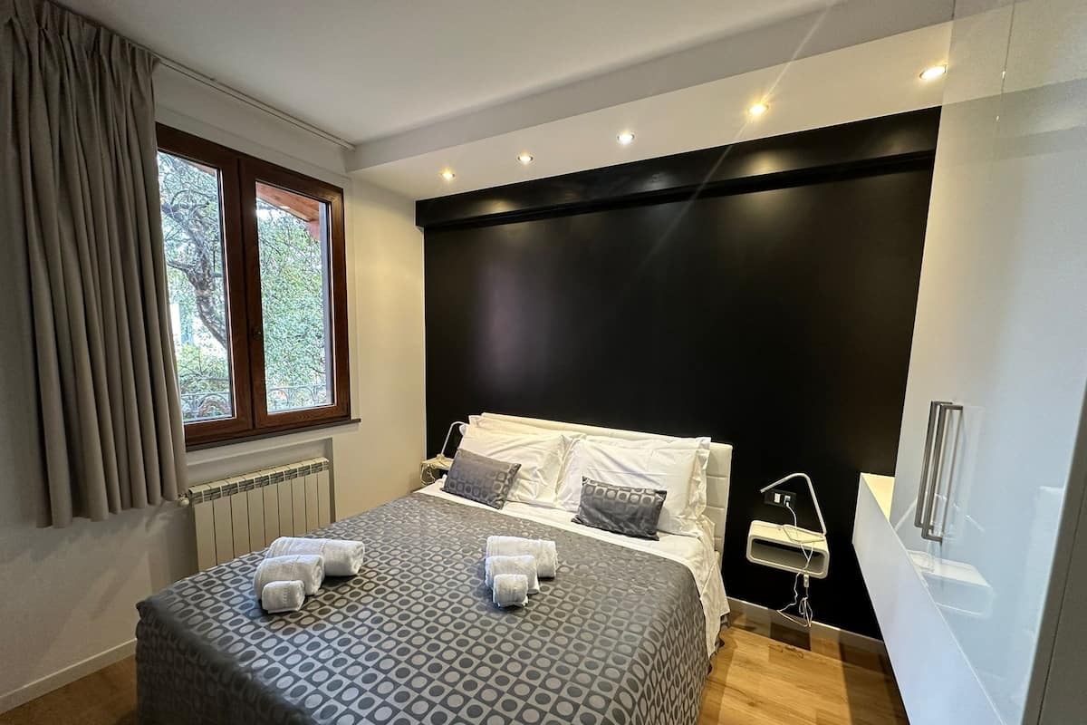 A modern bedroom with a large bed, a window with curtains, a black accent wall, and a bedside table with a lamp.