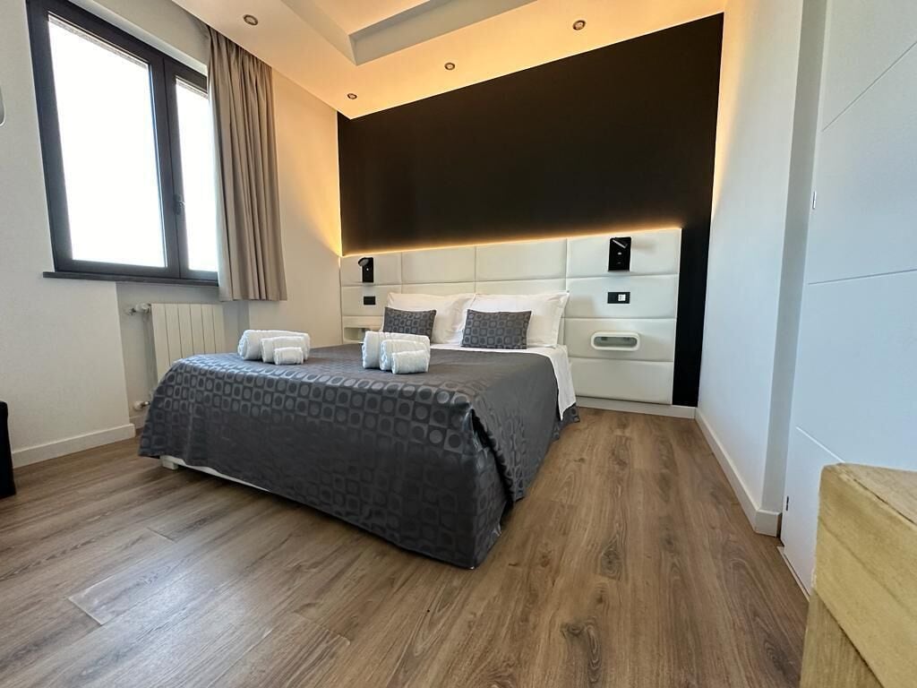 A modern bedroom with a large bed, a headboard with built-in lighting, and a window with curtains.