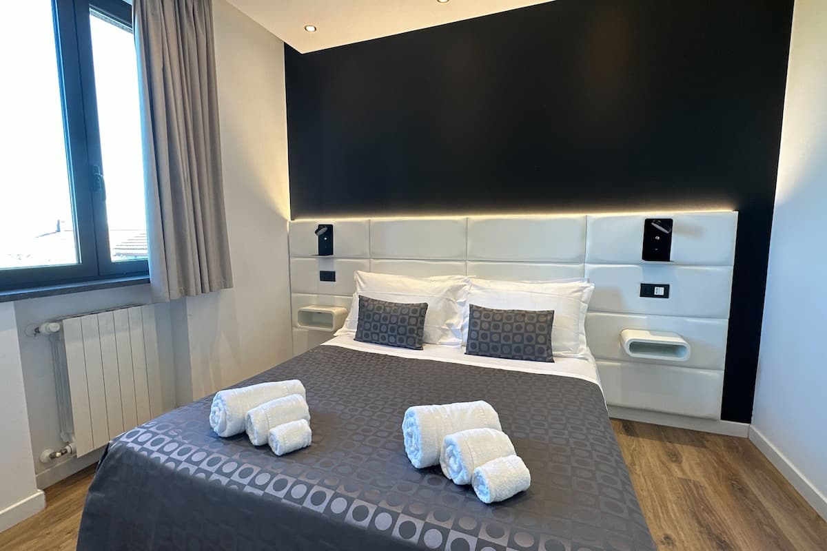 A modern hotel room with a large bed, a headboard with built-in lighting, a window with curtains, and a radiator.