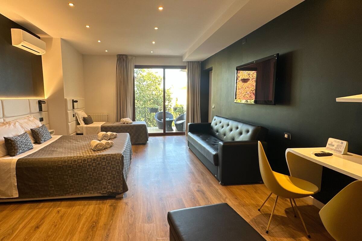 A modern hotel room with a large bed, a sofa, a desk, and a flat-screen TV.