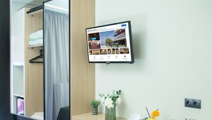 Et hotelværelse med en seng, et skrivebord med en computerskærm, der viser et hotelwebsted, en lille plante, en vase med blomster og et bakke med en kop og et glas appelsinjuice.