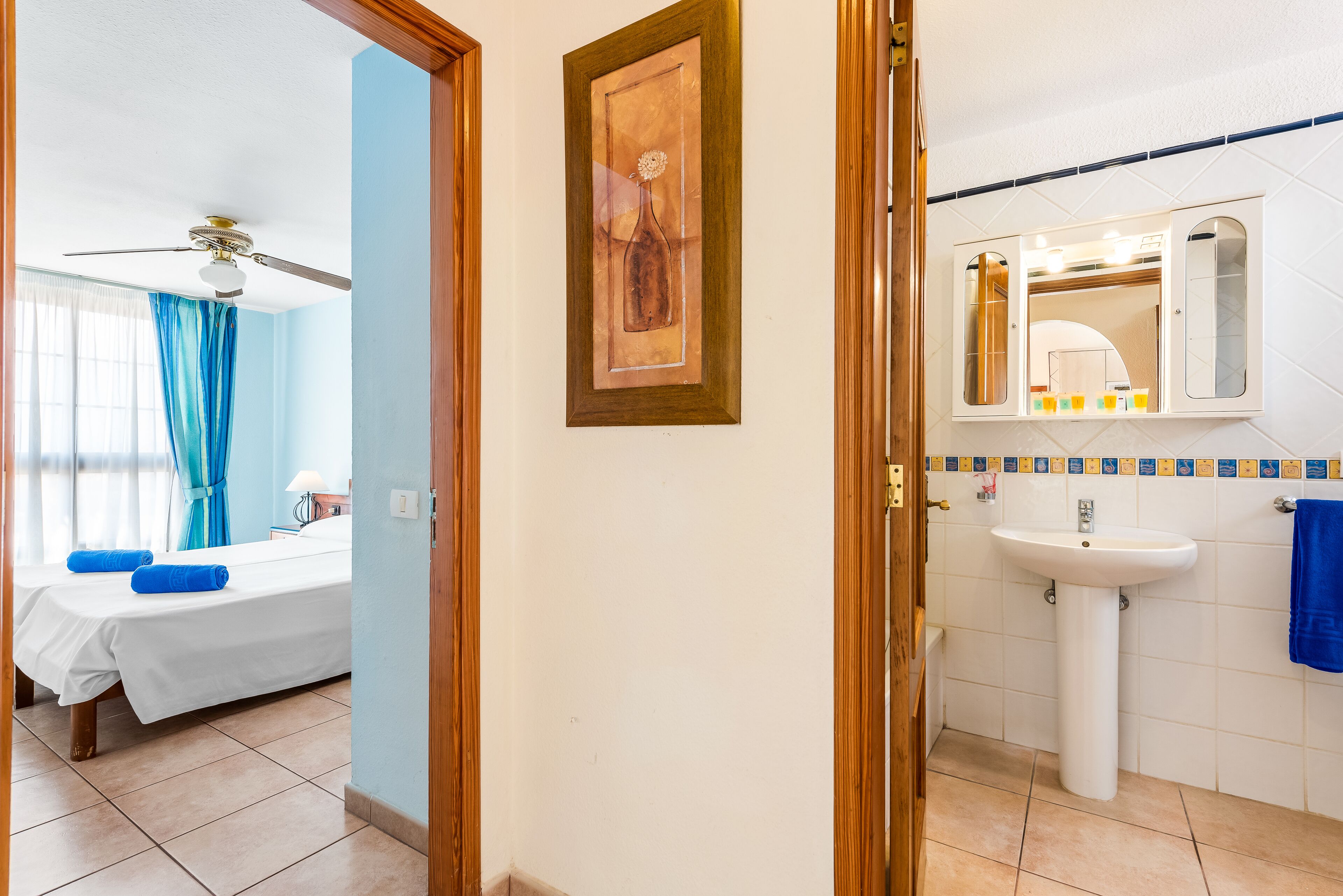 A hotel room with a bed, a ceiling fan, a painting, a bathroom with a sink and mirror.