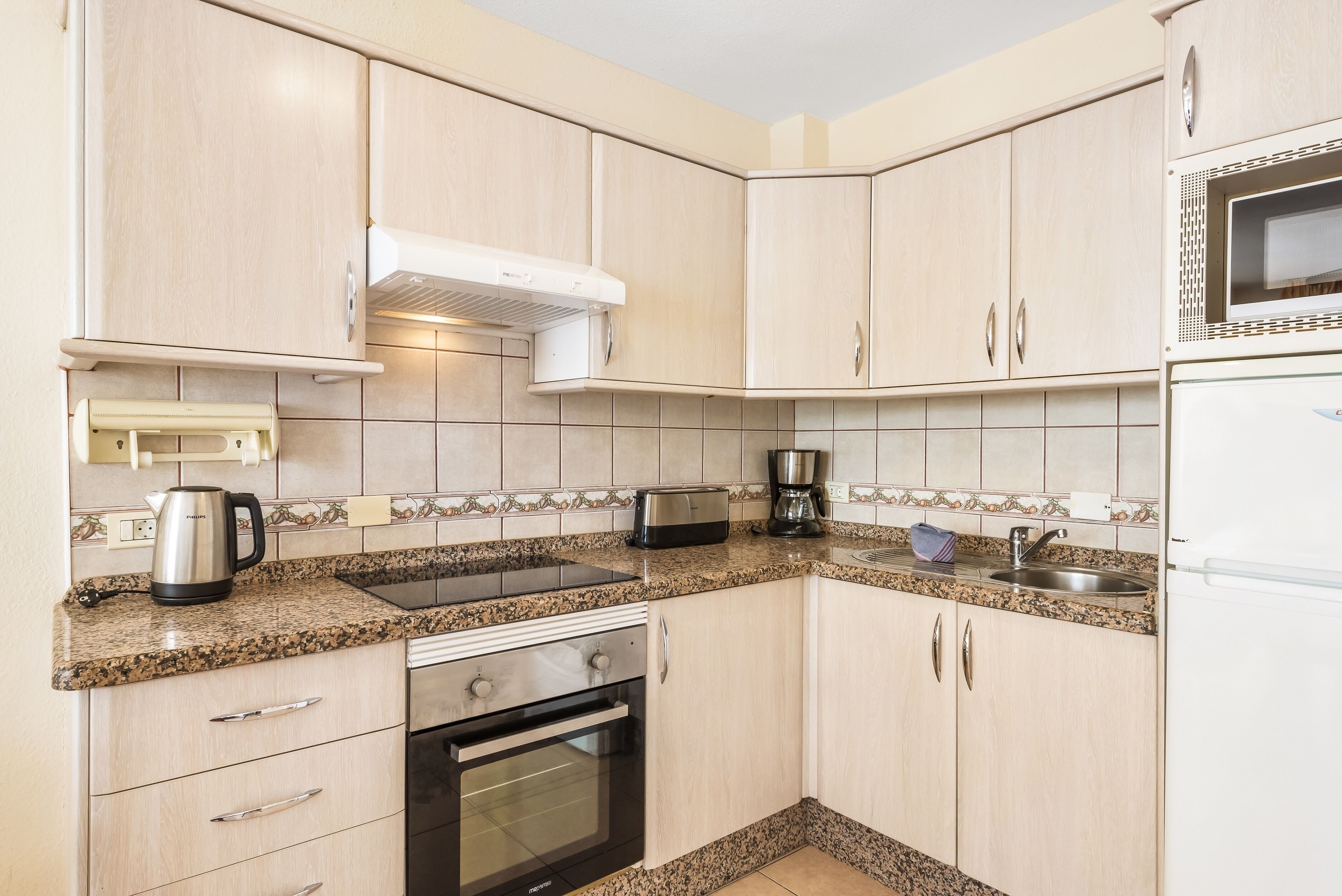A kitchen with light wood cabinets, a built-in oven, a microwave, a toaster, and a kettle on the countertop.
