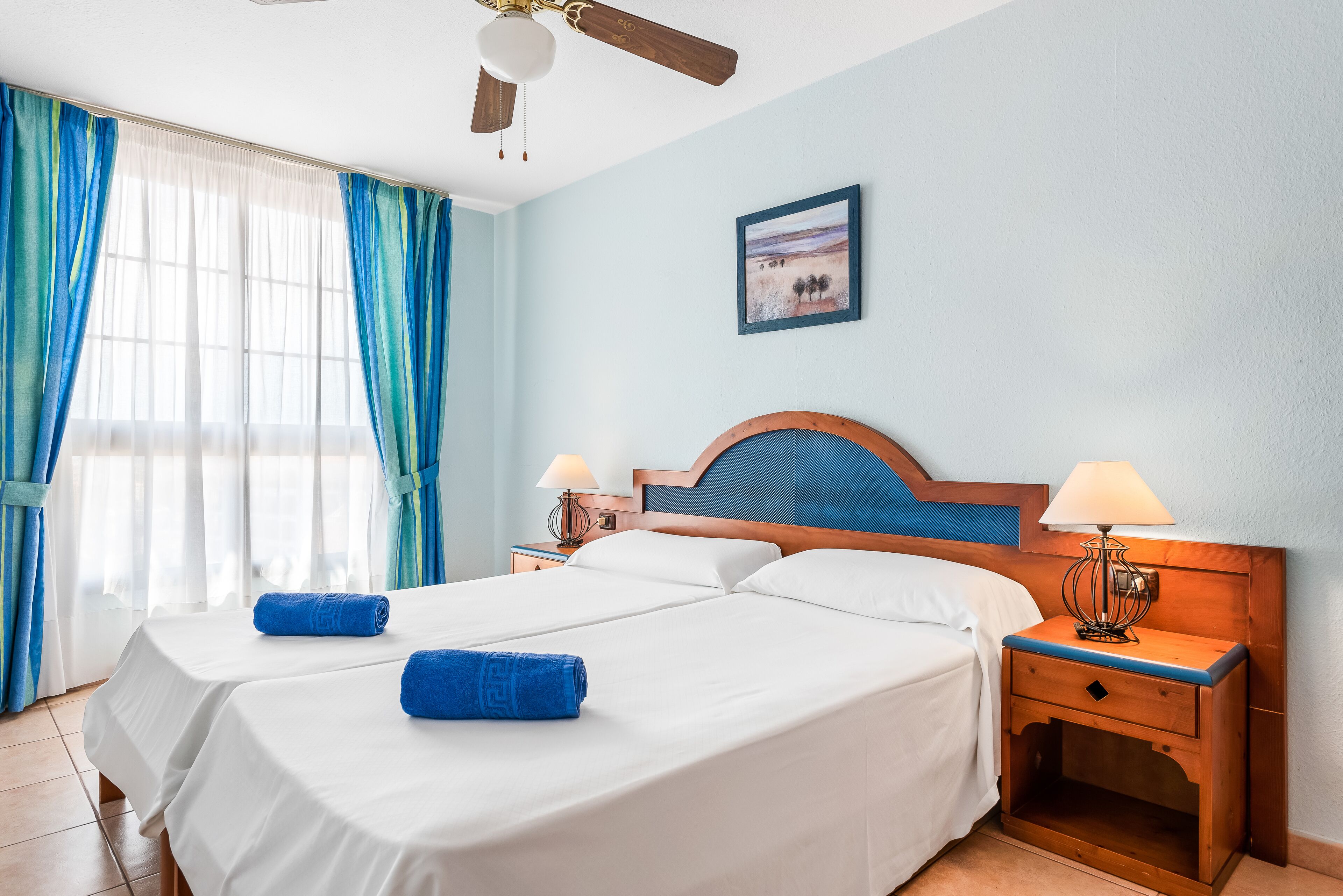 A hotel room with two beds, a ceiling fan, a nightstand with a lamp, and a painting on the wall.