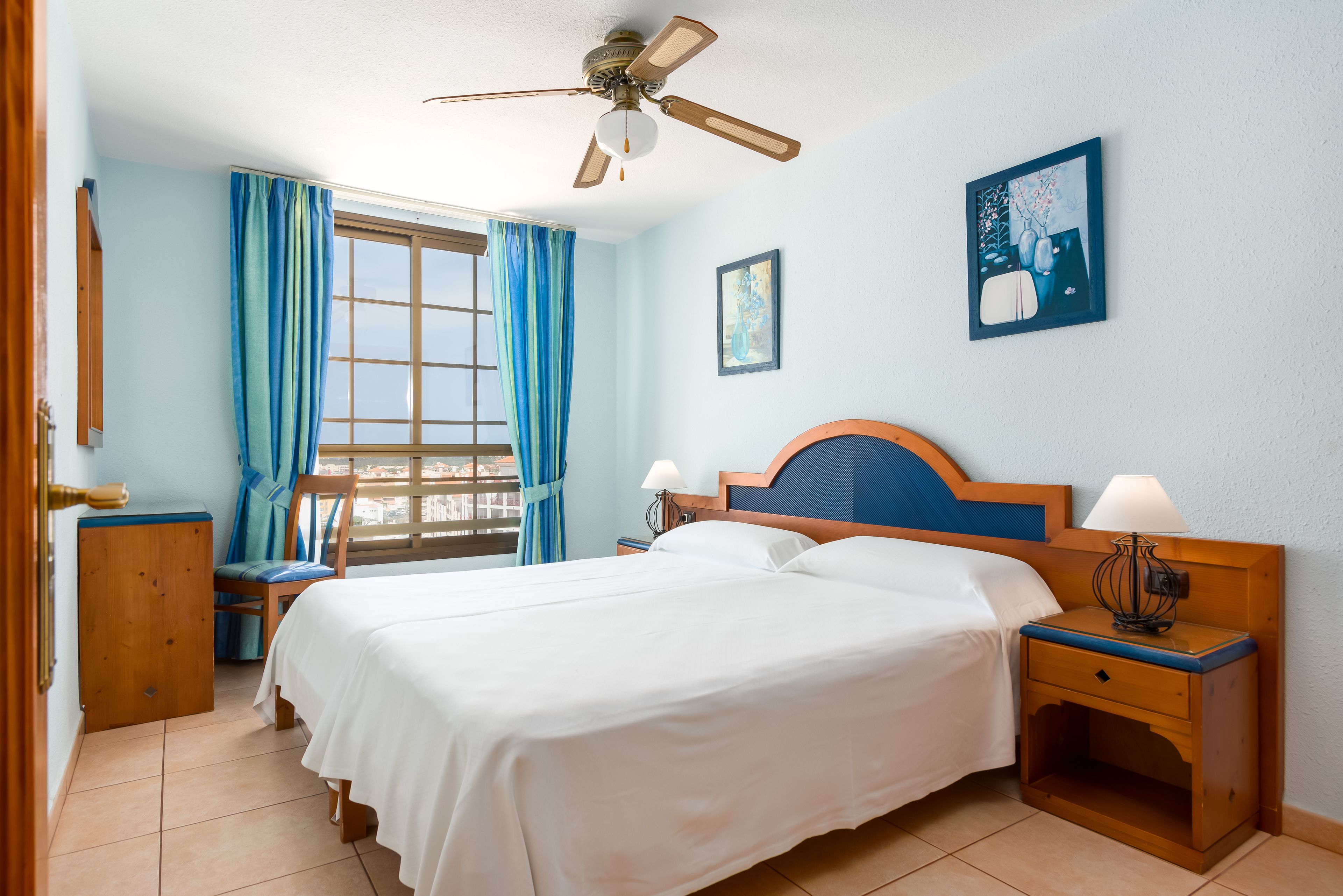 Ein Hotelzimmer mit Bett, Nachttisch, Schreibtisch, Stuhl, einem Fenster mit Vorhängen, einem Deckenventilator und Bildern an den Wänden.