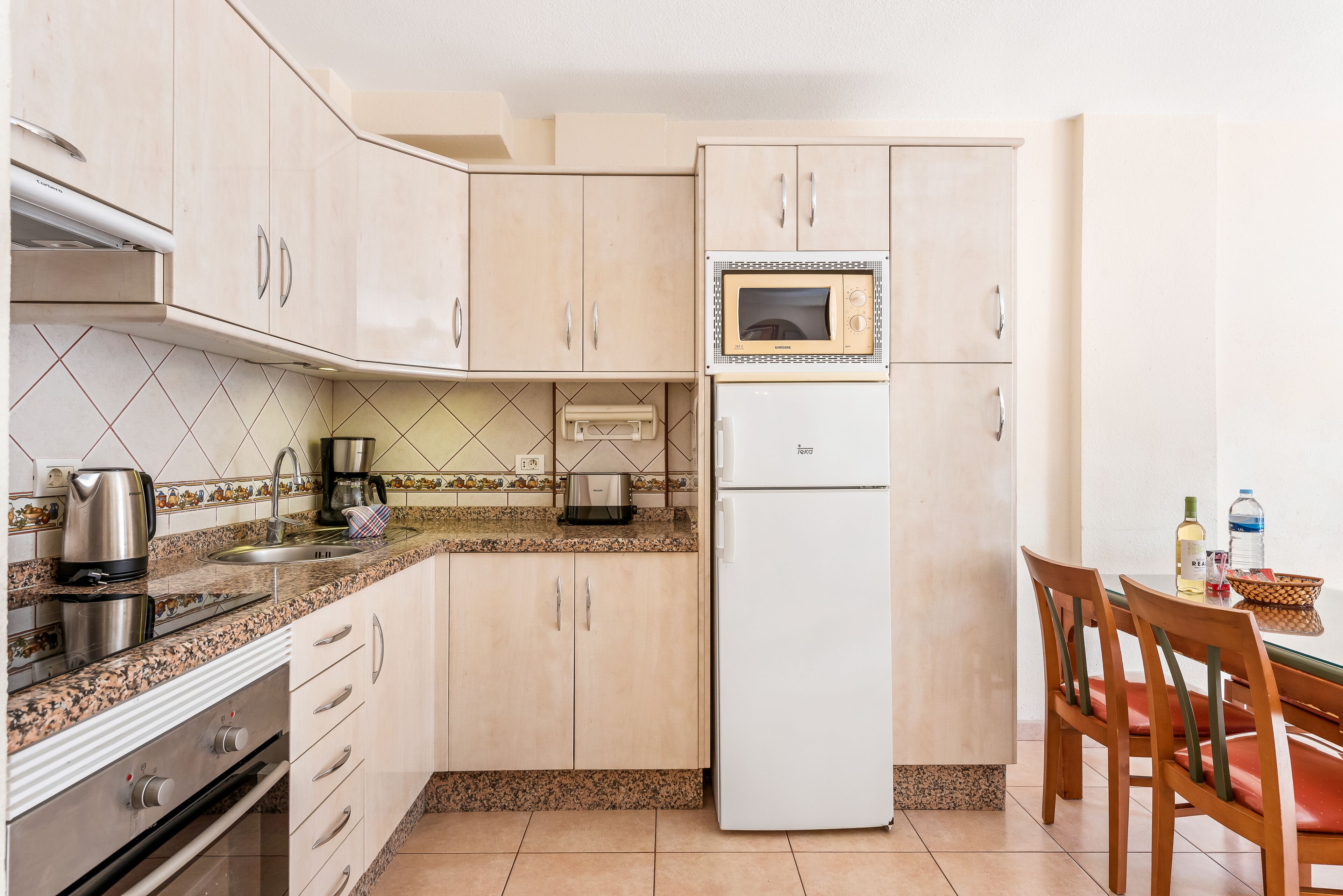 A compact kitchen with a microwave, stove, and refrigerator. There is a dining table with chairs and a coffee maker on the counter.