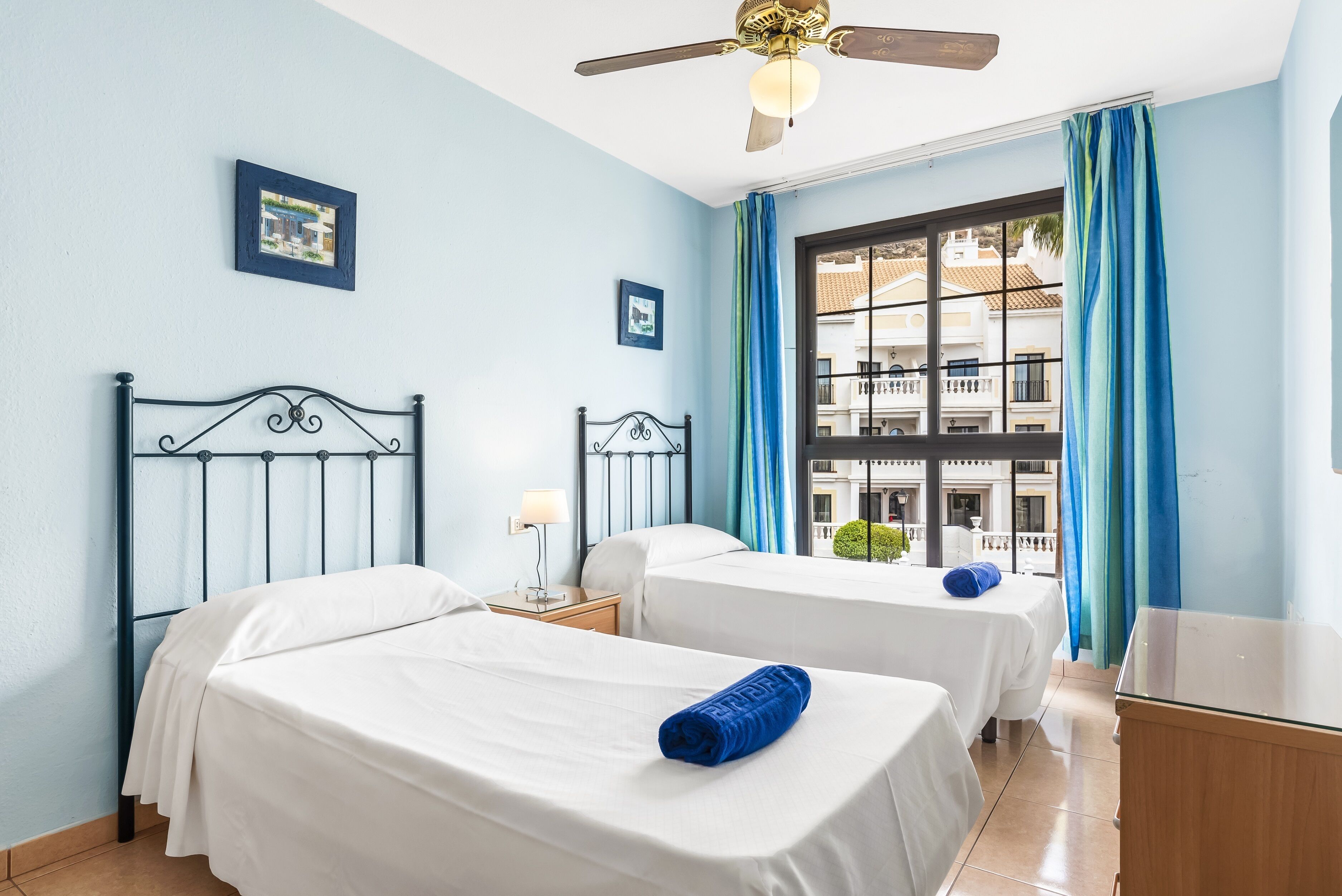 A hotel room with two beds, a ceiling fan, a window with blue curtains, and a view of buildings outside.