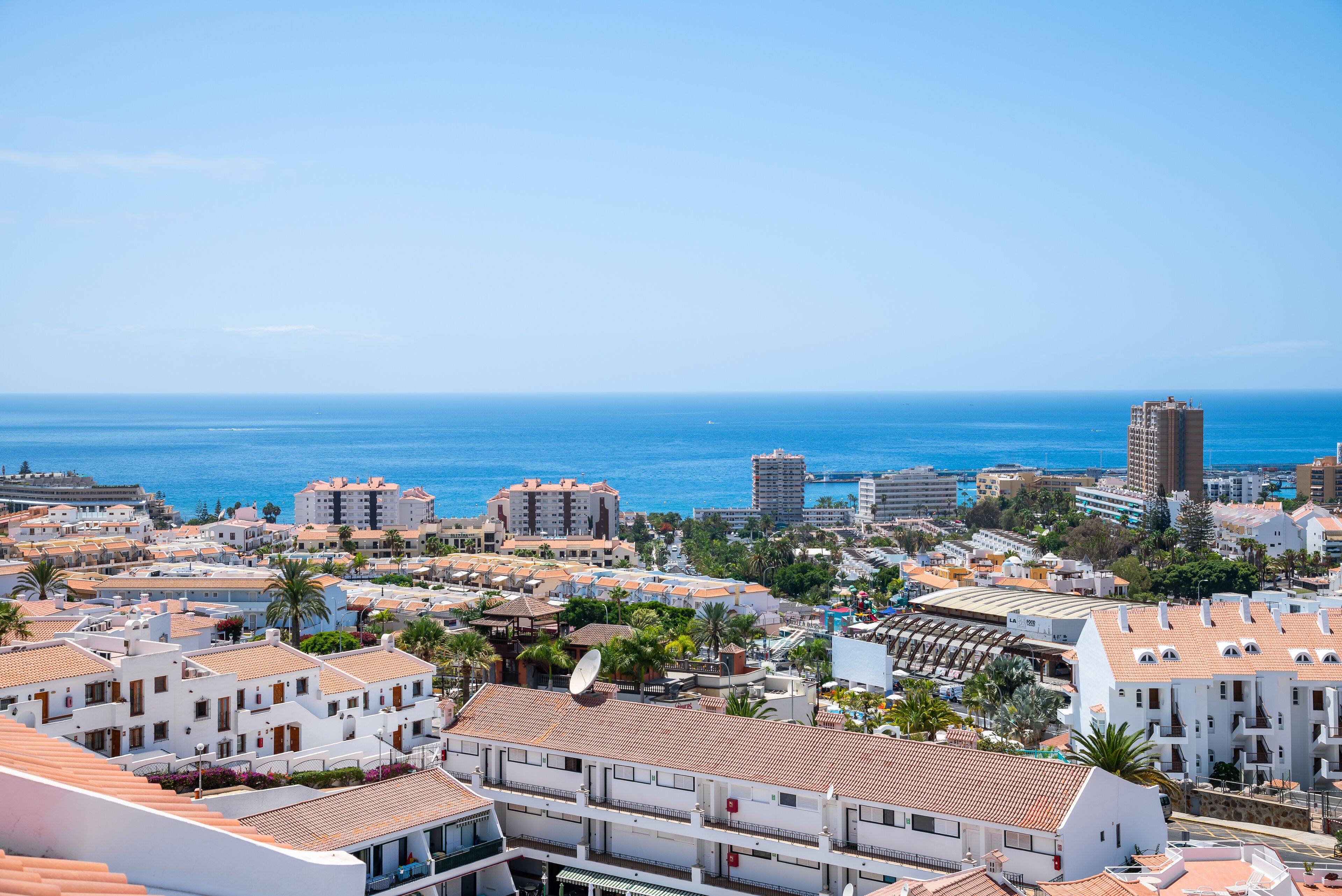 A coastal cityscape with white buildings, a clear blue sky, and the ocean in the background.