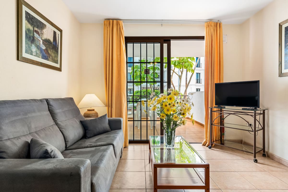 Un salon comprenant un canapé, une table basse en verre, un téléviseur posé sur un meuble, et une vue sur un balcon agrémenté de plantes.