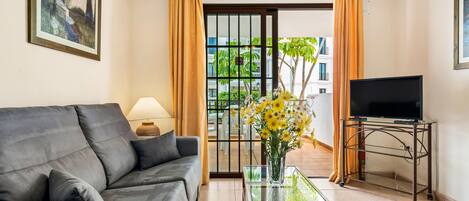 A living room with a sofa, a glass coffee table, a TV on a stand, and a view of a balcony with plants.