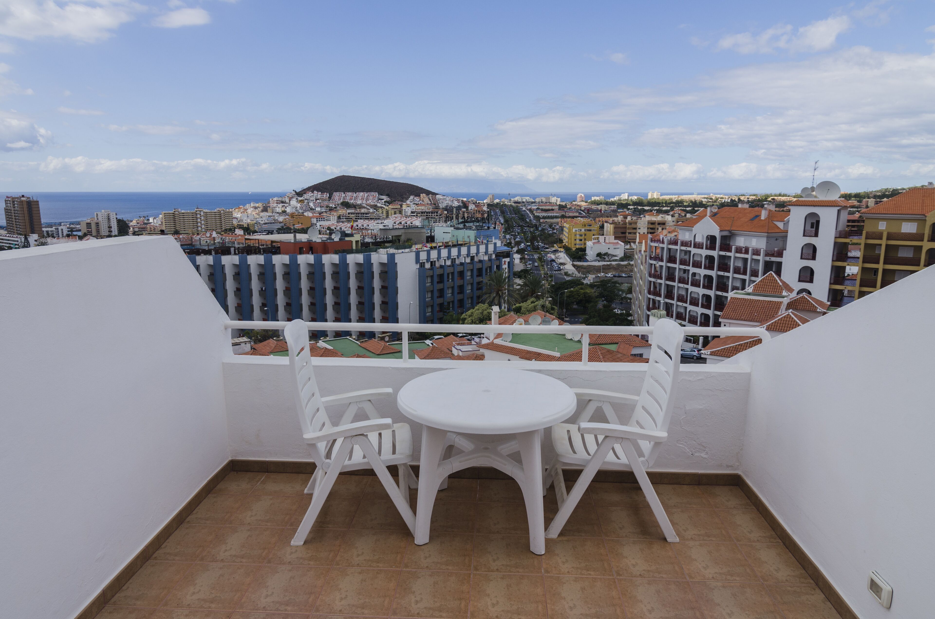 A balcony with a white table and chairs, overlooking a cityscape with buildings and a hill in the distance.