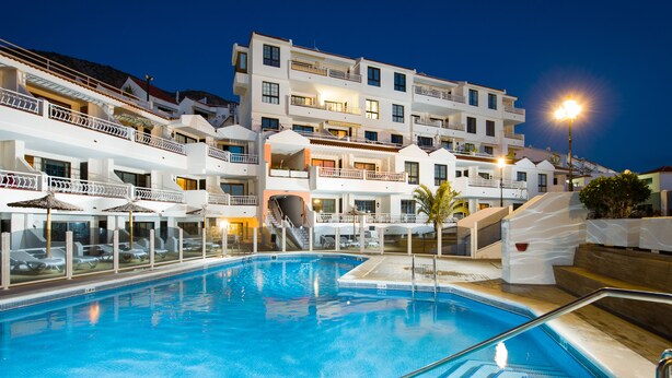 A swimming pool with a clear blue water, surrounded by white buildings with balconies and a staircase leading to the upper floors.