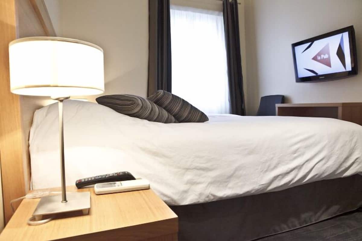 Una habitación de hotel con cama, mesita de noche con lámpara, control remoto y televisión en la pared.
