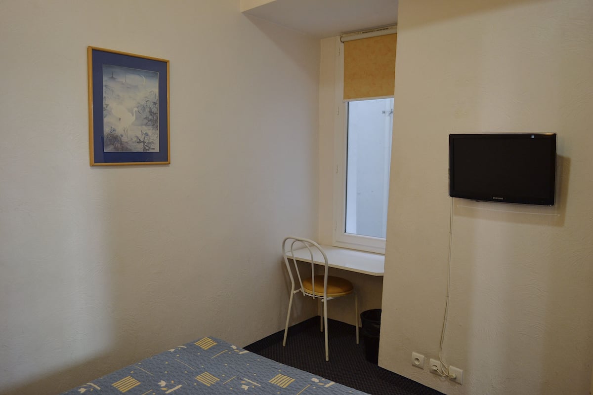 Una habitación pequeña con una cama, un escritorio con una silla, una ventana y un televisor de pantalla plana fijado a la pared.