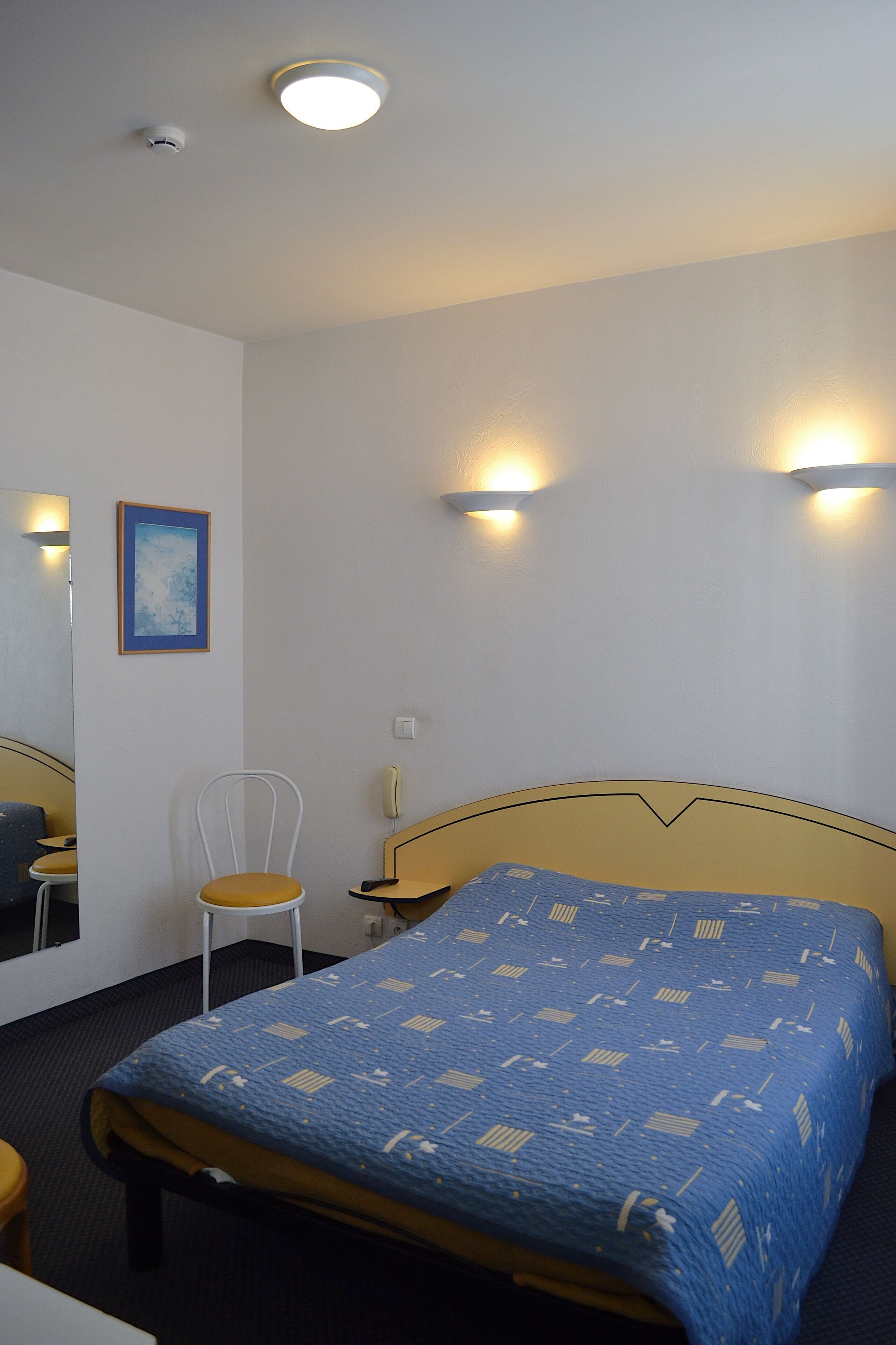 Une chambre d’hôtel comprenant un lit, une chaise, une table de chevet, un miroir et des lampes fixées au mur.
