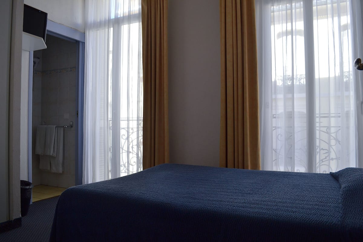 Habitación de hotel con cama, zona de ducha y balcón con cortinas.