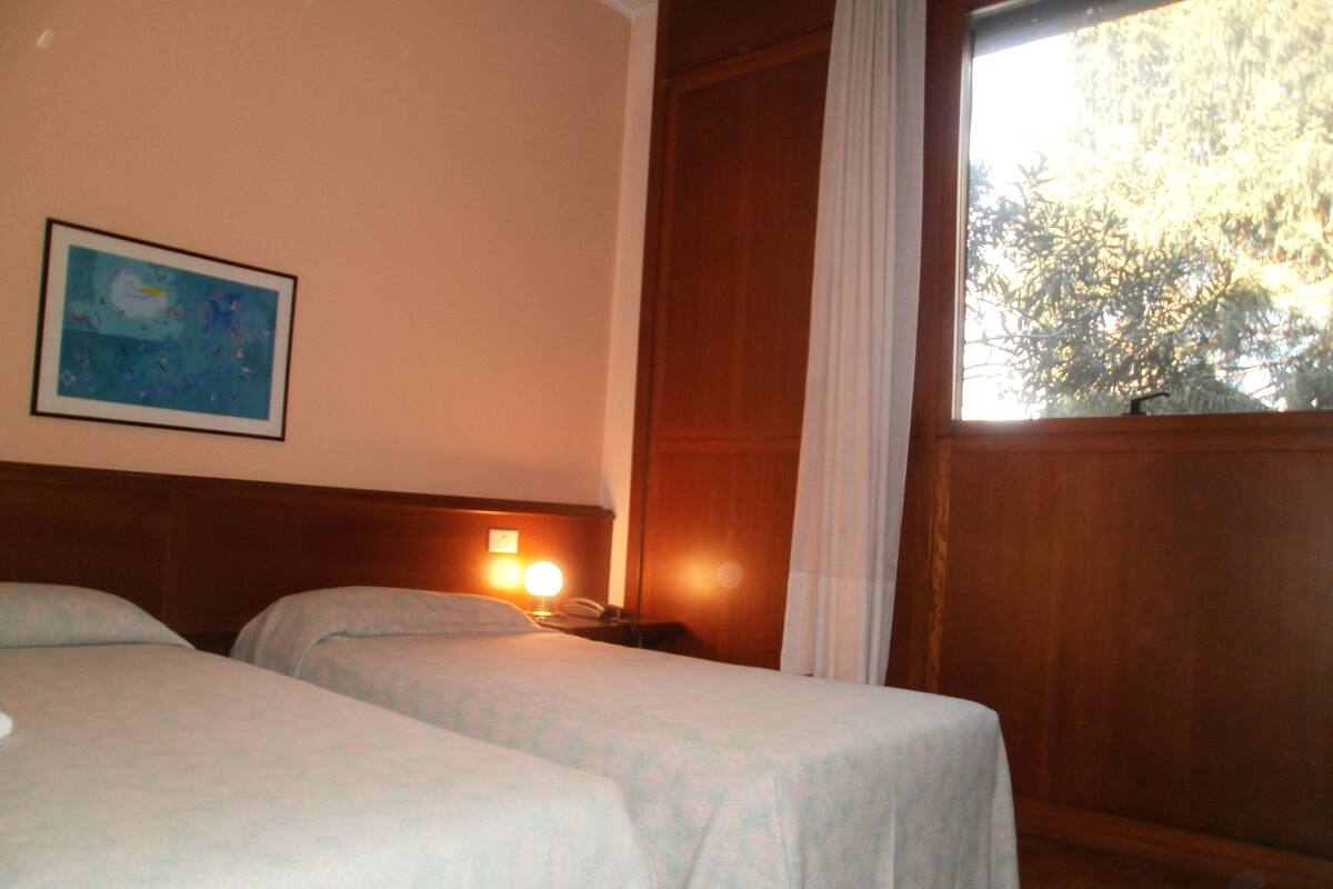 Una habitación con dos camas, un cuadro en la pared, una ventana con cortinas y una puerta de madera.