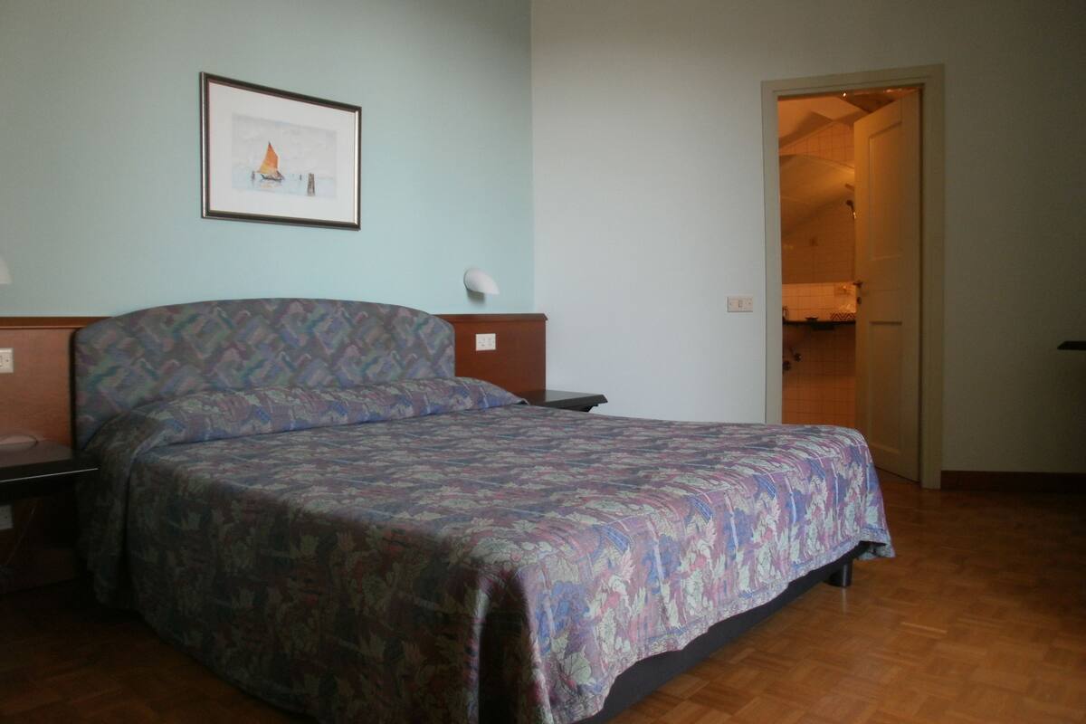 Un dormitorio con cama, mesita de noche, un cuadro enmarcado en la pared y una puerta que da al baño.