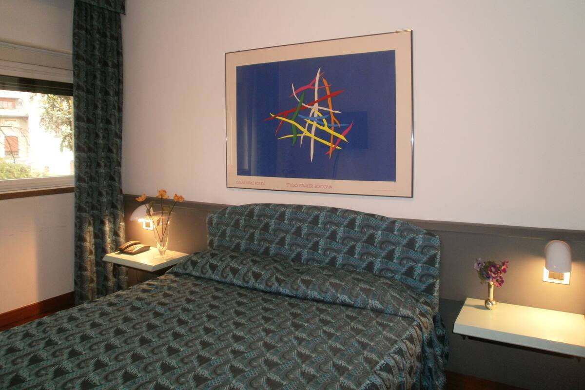 Un dormitorio con una colcha estampada, un cuadro abstracto enmarcado, una mesita de noche con una lámpara y una ventana con cortinas.