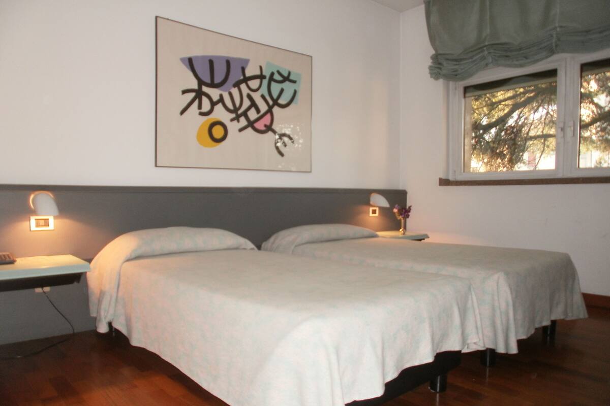 Un dormitorio con dos camas, una mesita de noche con lámpara, un ventanal con vistas a árboles y una pared decorada con una obra de arte abstracto.