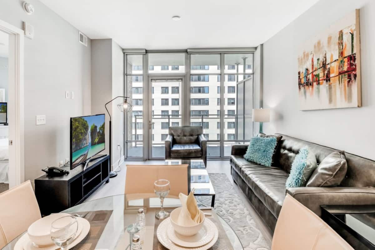 A modern hotel room with a dining area, a sofa, a television, and a large window with a city view.