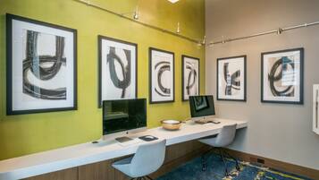 A modern office space with a long white desk, two computer monitors, a keyboard, and a mouse. The wall features several framed abstract art pieces.