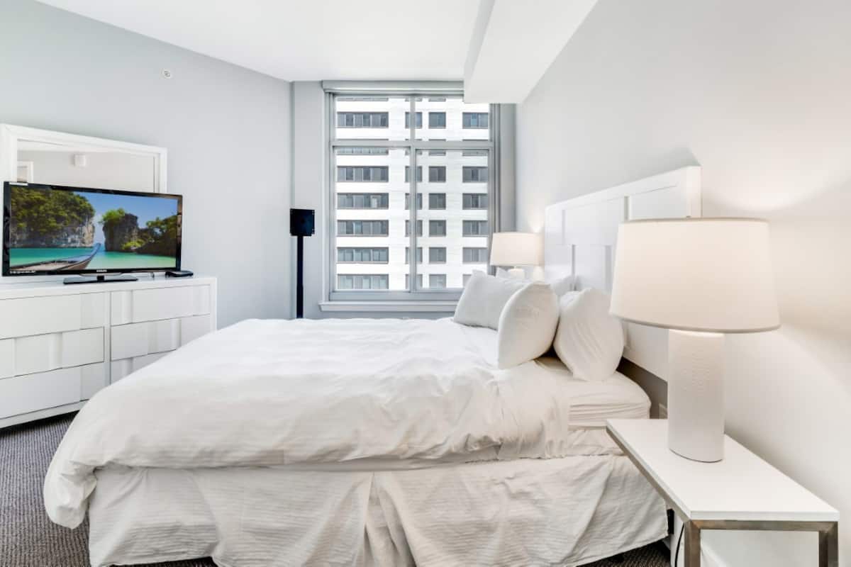 A hotel room with a bed, a TV, a window with a city view, and a lamp.