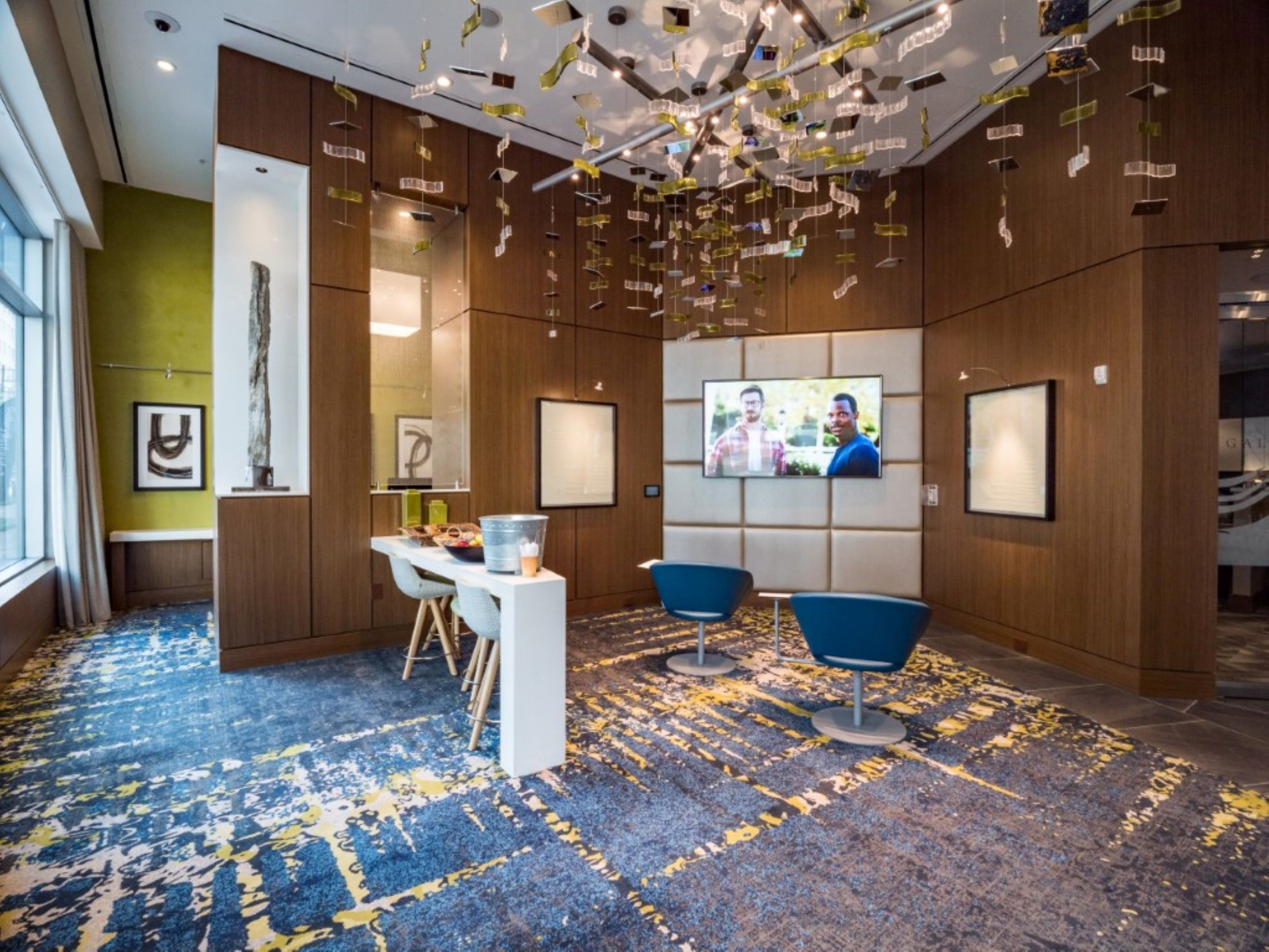 Eine moderne Lobby mit einem blau-gelb gemusterten Teppich, Holzpaneelen, einem Empfangstresen, zwei blauen Stühlen und einem an der Wand montierten Fernseher.