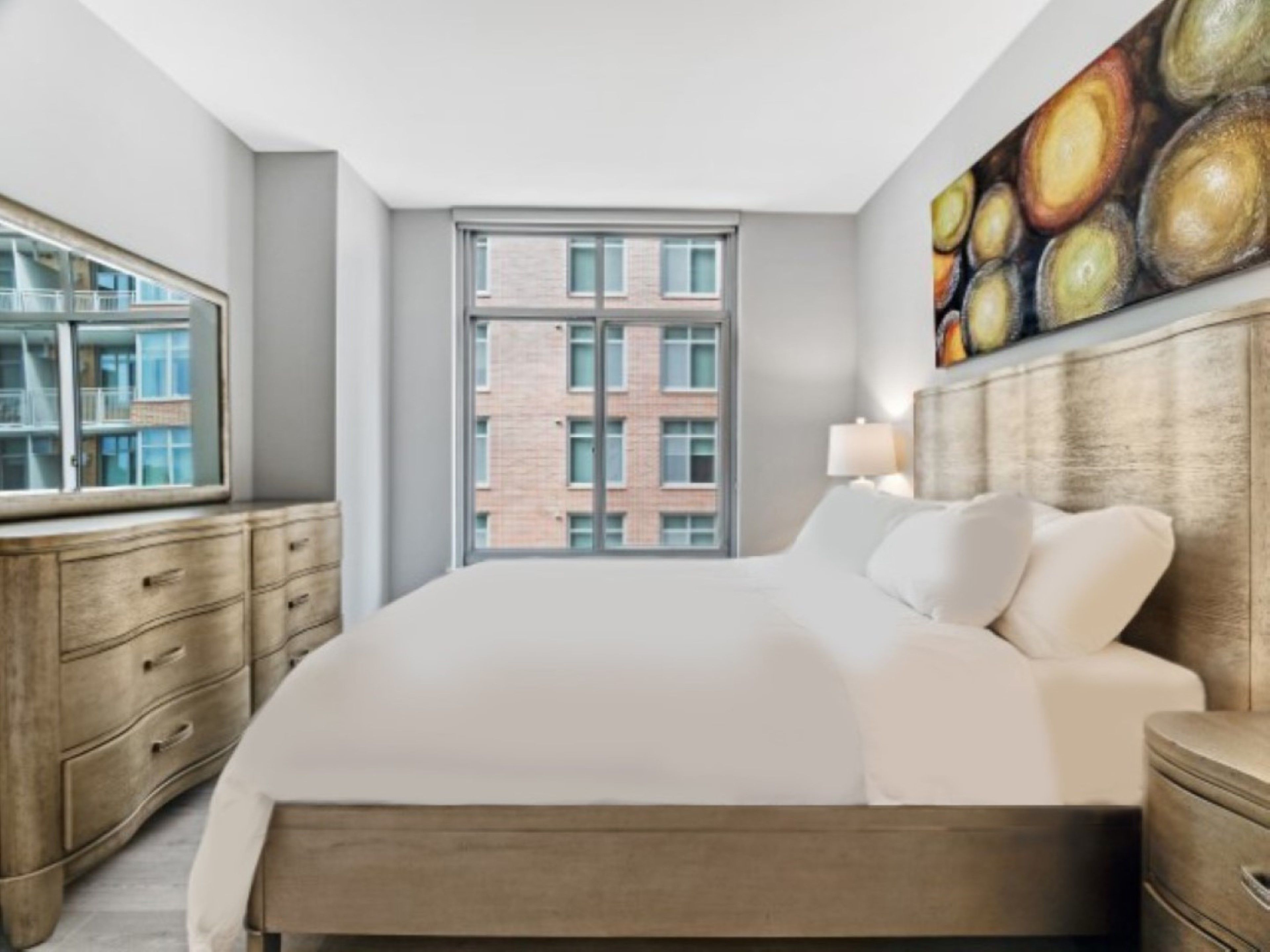 A modern hotel room with a large bed, a wooden dresser, a window with a city view, and a painting on the wall.