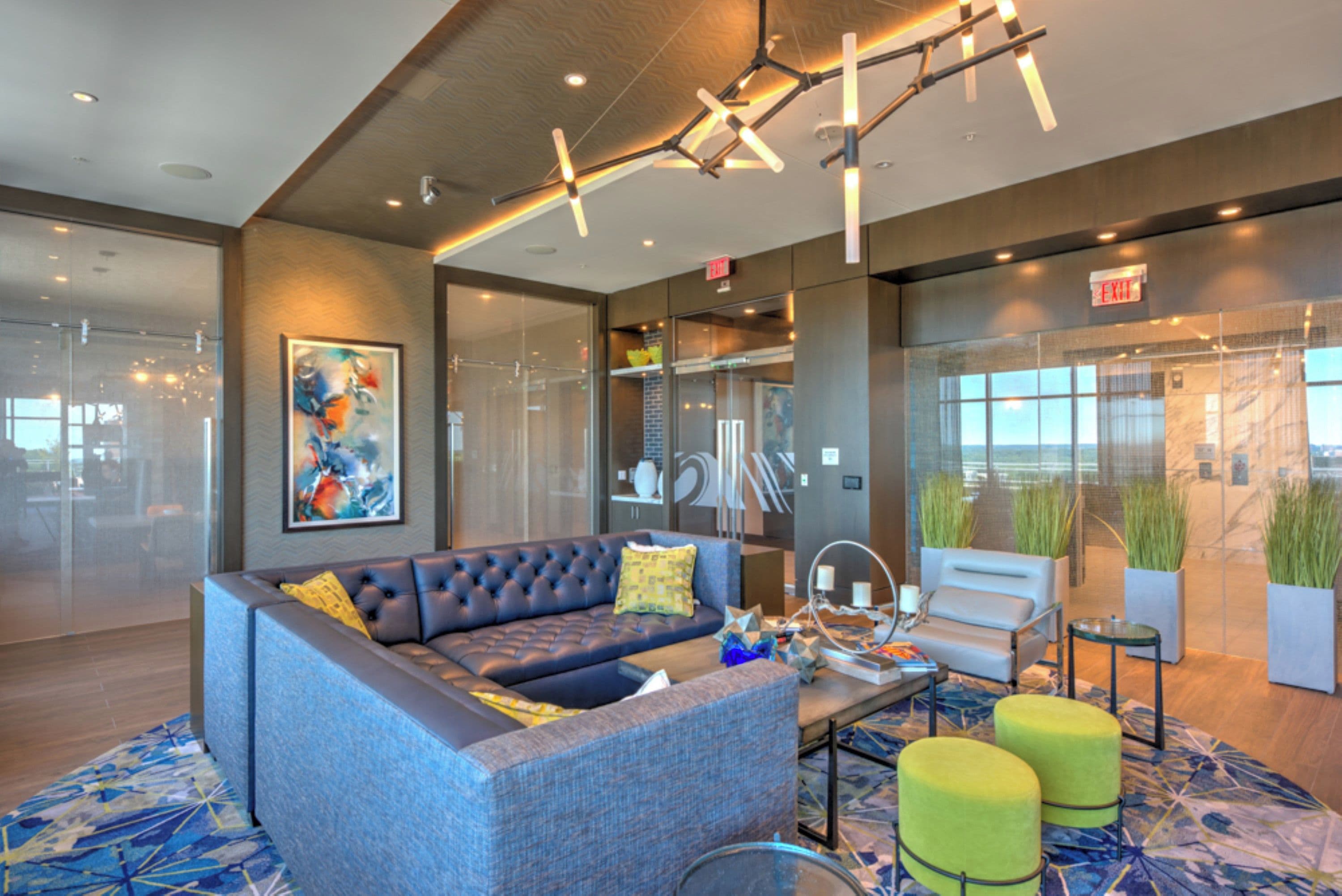 A modern lobby with a sectional sofa, a coffee table, and green ottomans. There is a glass wall with a view, and a painting on the wall.